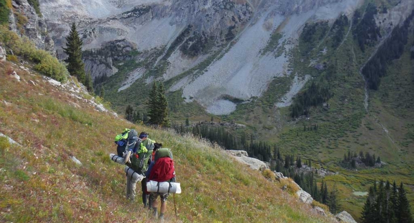 a group of students wearing backpacks hike across a grass field high above a mountainous valley